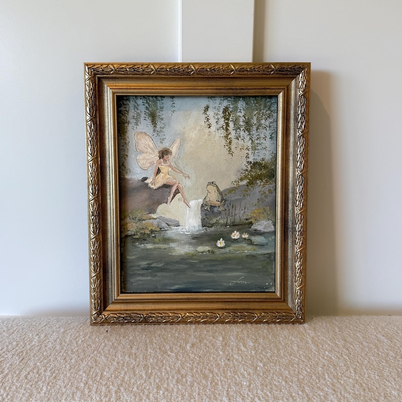 May include: A framed painting depicts a fairy with butterfly wings sitting near a waterfall, with a frog nearby. The artwork features a gold frame and a serene, whimsical scene with soft colors and details.