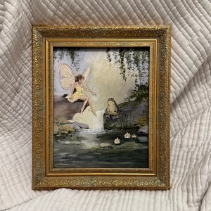 May include: A framed painting depicts a fairy in a yellow dress with large wings, seated near a waterfall. The artwork features a gold ornate frame and a serene landscape with water lilies and lush greenery. The painting evokes a whimsical, fantasy theme.