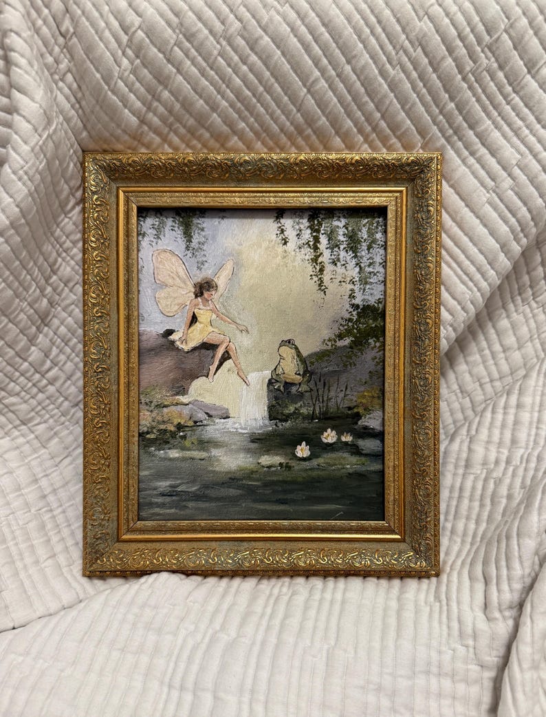 May include: A framed painting depicts a fairy in a yellow dress with large wings, seated near a waterfall. The artwork features a gold ornate frame and a serene landscape with water lilies and lush greenery. The painting evokes a whimsical, fantasy theme.