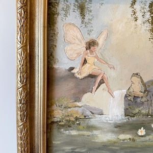 May include: A framed painting depicts a fairy in a yellow dress with large wings, seated near a waterfall. A green frog sits nearby. The artwork is set in a natural landscape with a gold-colored ornate frame.
