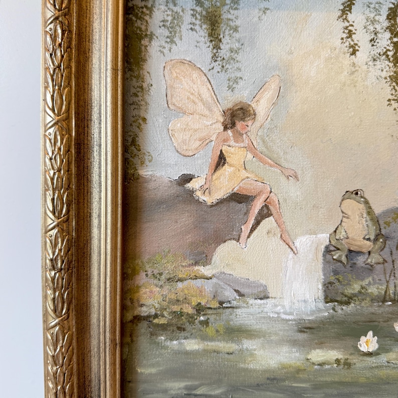 May include: A framed painting depicts a fairy in a yellow dress with large wings, seated near a waterfall. A green frog sits nearby. The artwork is set in a natural landscape with a gold-colored ornate frame.