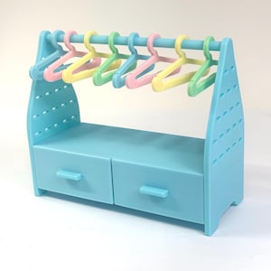 Peut inclure: Un portant à vêtements en plastique bleu clair doté de deux tiroirs de rangement à la base et d'une barre horizontale sur le dessus. Sept petits cintres aux couleurs pastel, rose, jaune et vert, sont suspendus à la barre supérieure.