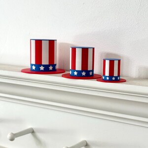 May include: Three decorative top hats in the style of Uncle Sam. Each hat has red and white stripes, a blue brim, and a band of white stars. The hats are displayed on a white shelf.