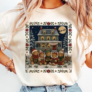 Puede incluir: Una camiseta de manga corta color crema con un gráfico rectangular de una casa de dos pisos de noche con luna llena, árboles nevados y cinco osos de peluche con gorros y bufandas de invierno, cada uno sosteniendo un regalo, con borde de acebo.