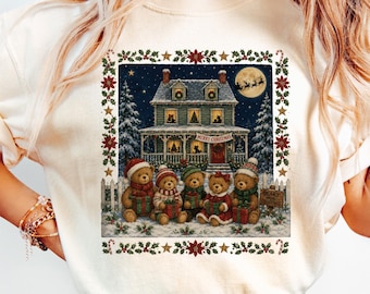 Puede incluir: Una camiseta de manga corta color crema con un gráfico rectangular de una casa de dos pisos de noche con luna llena, árboles nevados y cinco osos de peluche con gorros y bufandas de invierno, cada uno sosteniendo un regalo, con borde de acebo.