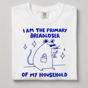 May include: A white t-shirt featuring a blue graphic of a mouse wearing sunglasses, holding a martini glass and a bag of bread. The text reads, I AM THE PRIMARY BREADLOSER OF MY HOUSEHOLD. The shirt has a Comfort Colors brand label at the neck.