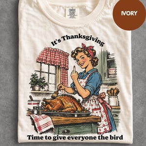 Puede incluir: Una camiseta de color marfil con una ilustración gráfica de una persona en una cocina preparando un pavo asado. El texto dice It's Thanksgiving y Time to give everyone the bird. La persona viste una camisa azul y un delantal estampado.