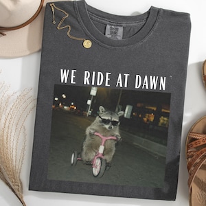 May include: Dark gray Comfort Colors t-shirt with the text "WE RIDE AT DAWN" above an image of a raccoon wearing sunglasses and riding a pink tricycle. A gold necklace and a beige hat are also visible.