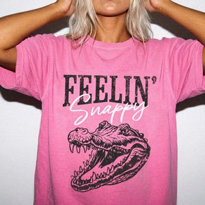 May include: A bright pink short-sleeve t-shirt featuring a black graphic of an alligator head with its mouth open. The text "FEELIN'" is printed in a distressed black font above the word "Snappy" written in a white cursive script.