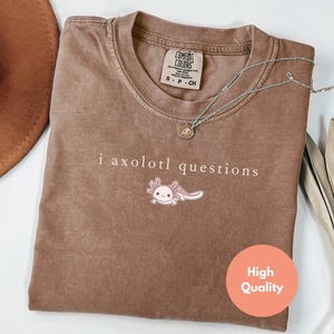 May include: A brown Comfort Colors t-shirt featuring the text "i axolotl questions" and an axolotl cartoon. A silver necklace with a pendant is on the shirt. A brown hat is visible in the background.