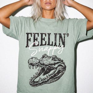 May include: A sage green short-sleeve t-shirt featuring a black distressed graphic of an alligator with its mouth open. The text "FEELIN'" is printed in a bold, distressed black font above the word "Snappy" in a white cursive script.