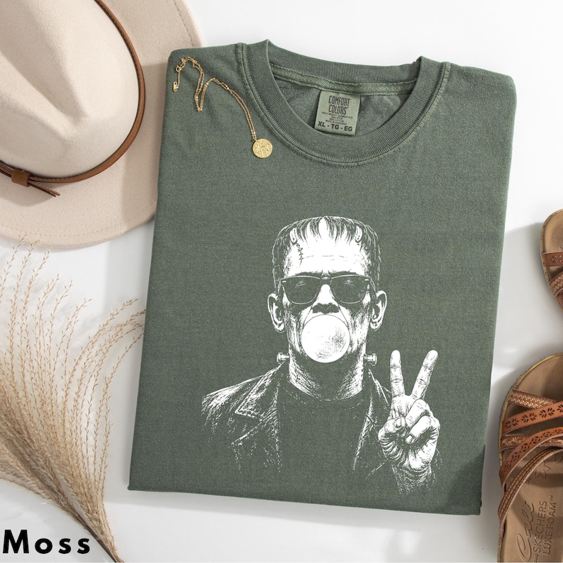 Puede incluir: Una camiseta verde musgo con un estampado gr&aacute;fico blanco de un personaje con corte de pelo plano, gafas de sol, inflando un chicle y haciendo el signo de la paz con una mano. La camiseta tiene cuello redondo y una etiqueta interior.