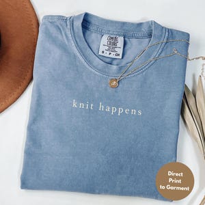 May include: A blue Comfort Colors t-shirt with the phrase "knit happens" embroidered in white. A gold necklace with a pendant rests on the shirt. The shirt is folded, and a brown hat is visible in the background.