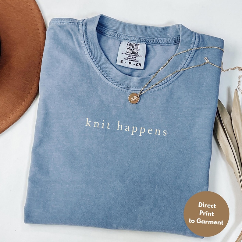 May include: A blue Comfort Colors t-shirt with the phrase "knit happens" embroidered in white. A gold necklace with a pendant rests on the shirt. The shirt is folded, and a brown hat is visible in the background.