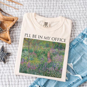 May include: A beige t-shirt with the text "I'll be in my office" printed on the front. The text is in black and is centered on the shirt. The shirt is folded and laid on a white surface. A painting of a garden with flowers and trees is printed on the shirt.