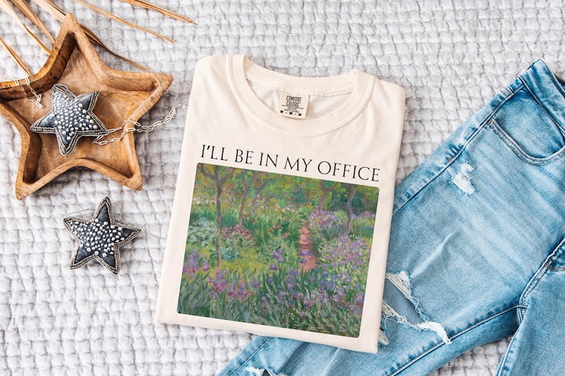 May include: A beige t-shirt with the text "I'll be in my office" printed on the front. The text is in black and is centered on the shirt. The shirt is folded and laid on a white surface. A painting of a garden with flowers and trees is printed on the shirt.