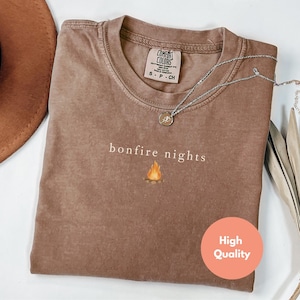 May include: A brown Comfort Colors t-shirt with the words "bonfire nights" and a small bonfire graphic. A silver necklace with a pendant is on the shirt. The shirt is folded and has a "High Quality" sticker.
