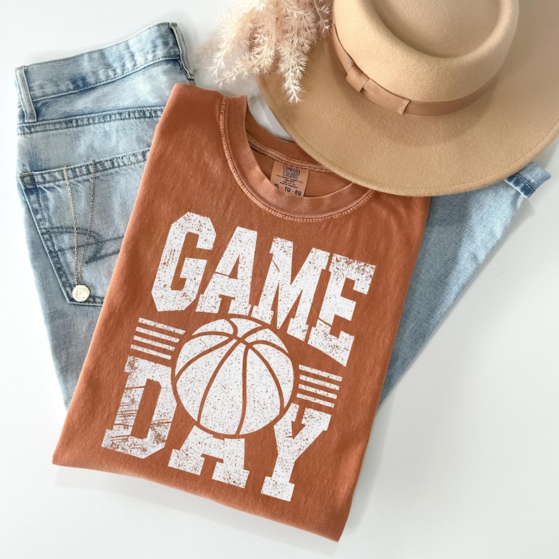 Puede incluir: Una camiseta naranja con las palabras "GAME DAY" y un gr&aacute;fico de baloncesto, combinada con vaqueros azul claro y un sombrero color beige. Se ve un collar dorado. La camiseta es de estilo casual, adecuada para los aficionados al deporte.
