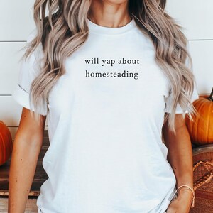 K&ouml;nnte beinhalten: Wei&szlig;es T-Shirt mit dem Text "will yap about homesteading" in schwarzer Schrift. Das T-Shirt mit Rundhalsausschnitt hat einen klassischen Stil. K&uuml;rbisse im Bild deuten auf ein Herbstthema hin.