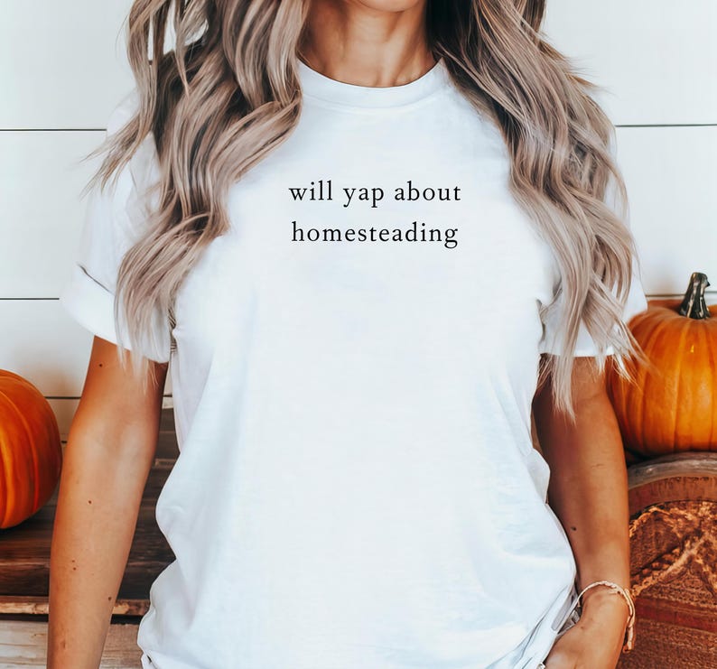 K&ouml;nnte beinhalten: Wei&szlig;es T-Shirt mit dem Text "will yap about homesteading" in schwarzer Schrift. Das T-Shirt mit Rundhalsausschnitt hat einen klassischen Stil. K&uuml;rbisse im Bild deuten auf ein Herbstthema hin.