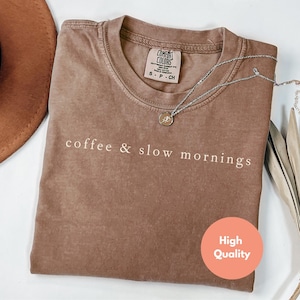 Może przedstawiać: Brązowa koszulka Comfort Colors z napisem "coffee & slow mornings" w białej czcionce. Koszulka ma okrągły dekolt i kr&oacute;tkie rękawy. Srebrny naszyjnik z wisiorkiem jest zarzucony na koszulkę. Brązowy kapelusz jest widoczny w tle.
