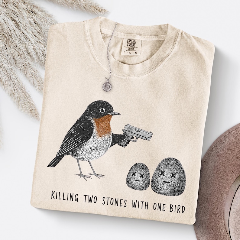 Puede incluir: Una camiseta beige con un gr&aacute;fico de un p&aacute;jaro sosteniendo una pistola y apuntando a dos piedras con ojos en forma de X. El texto debajo del gr&aacute;fico dice: "KILLING TWO STONES WITH ONE BIRD." La camiseta tiene una etiqueta de marca Comfort Colors.