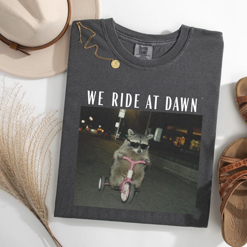 May include: Dark gray Comfort Colors t-shirt with the text "WE RIDE AT DAWN" above an image of a raccoon wearing sunglasses and riding a pink tricycle. A gold necklace and a beige hat are also visible.