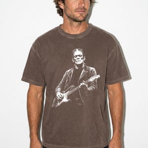 Puede incluir: Una camiseta marr&oacute;n de manga corta con un estampado gr&aacute;fico blanco de una figura que se asemeja al monstruo de Frankenstein, vistiendo una chaqueta y tocando una guitarra el&eacute;ctrica. La camiseta tiene cuello redondo y un aspecto desgastado.