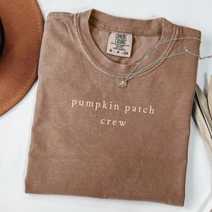 Puede incluir: Una camiseta marrón con las palabras "pumpkin patch crew" impresas en la parte delantera. La camiseta tiene cuello redondo y mangas cortas. Un collar plateado con un pequeño colgante está sobre la camiseta.
