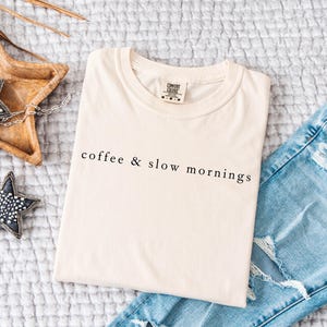 Może przedstawiać: Kremowa koszulka z czarnym napisem "coffee & slow mornings", złożona na teksturowanej powierzchni obok jasnoniebieskich podartych dżins&oacute;w. Obecne są r&oacute;wnież ozdoby w kształcie gwiazd i drewniana taca.