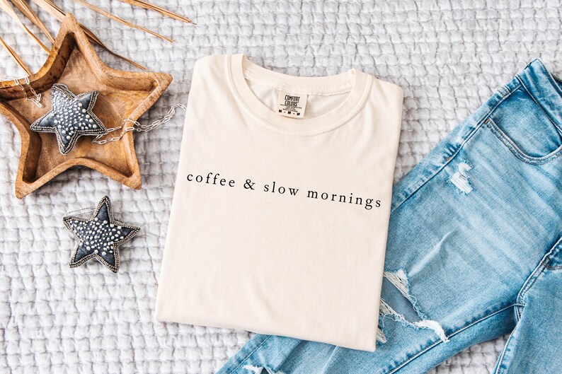 Może przedstawiać: Kremowa koszulka z czarnym napisem "coffee & slow mornings", złożona na teksturowanej powierzchni obok jasnoniebieskich podartych dżins&oacute;w. Obecne są r&oacute;wnież ozdoby w kształcie gwiazd i drewniana taca.