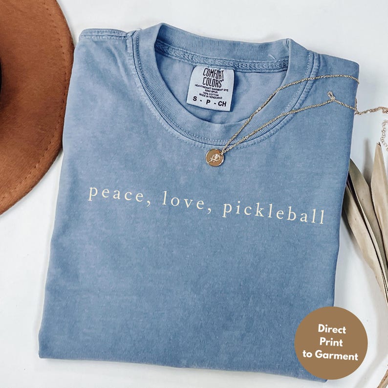 May include: A blue Comfort Colors t-shirt with the words "peace, love, pickleball" printed in white. A gold necklace with a pendant rests on the shirt. The shirt is folded, and a brown hat is visible in the background.