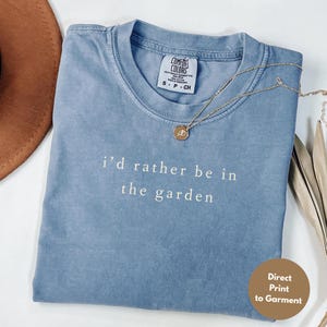 May include: A blue Comfort Colors t-shirt with the text "i'd rather be in the garden" in white. A gold necklace with a pendant rests on the shirt. A brown hat is partially visible in the background. The shirt is a size small.