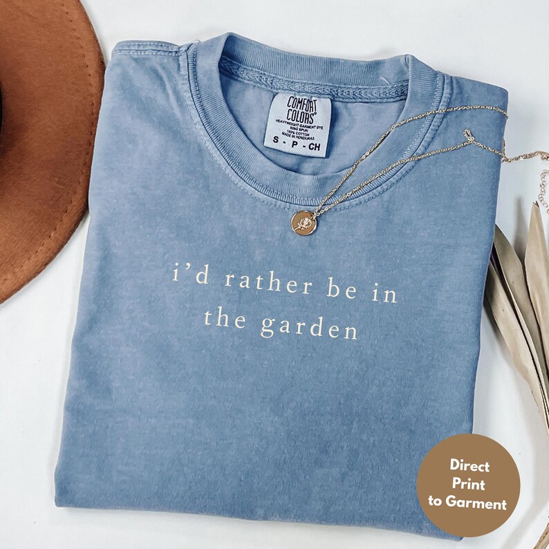 May include: A blue Comfort Colors t-shirt with the text "i'd rather be in the garden" in white. A gold necklace with a pendant rests on the shirt. A brown hat is partially visible in the background. The shirt is a size small.
