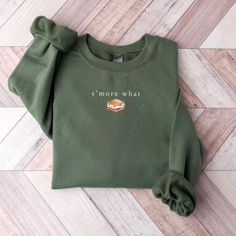 May include: Olive green sweatshirt with the text "s'more what" and a graphic of a s'more. The long-sleeved top is made of a soft material and is suitable for casual wear.