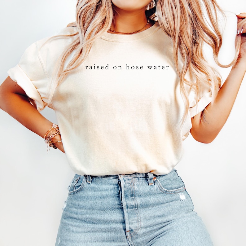 May include: Cream-colored t-shirt with the phrase "raised on hose water" printed in black. The shirt is paired with blue denim jeans. The t-shirt has rolled-up sleeves.