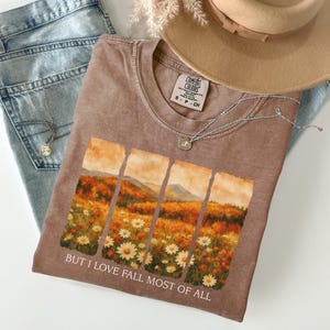 May include: A brown t-shirt with a landscape graphic featuring autumn colours and daisies. The text "BUT I LOVE FALL MOST OF ALL" is printed below the graphic. The shirt is paired with light wash jeans, a tan hat, and a silver necklace.