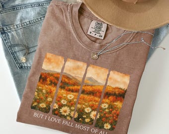 May include: A brown t-shirt with a landscape graphic featuring fall colors and daisies. The text "BUT I LOVE FALL MOST OF ALL" is printed below the graphic. The shirt is paired with light wash jeans, a tan hat, and a silver necklace.