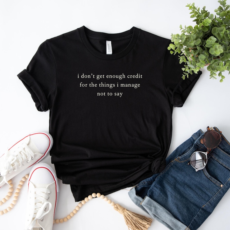 May include: Black t-shirt with the text "i don't get enough credit for the things i manage not to say" in a white, sans-serif font. The shirt is styled with white sneakers, denim shorts, and sunglasses.