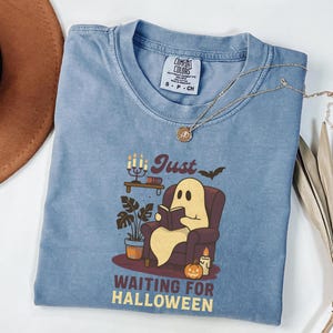 May include: A blue Comfort Colors t-shirt with a graphic of a ghost reading in a chair, a candle, and the text "Just Waiting for Halloween". A gold necklace and a brown hat are also in the image.