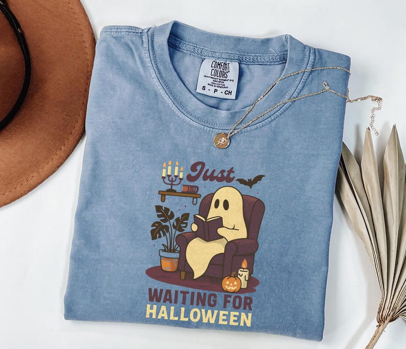 May include: A blue Comfort Colors t-shirt with a graphic of a ghost reading in a chair, a candle, and the text "Just Waiting for Halloween". A gold necklace and a brown hat are also in the image.