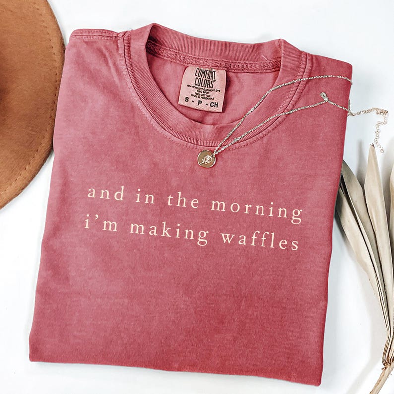 K&ouml;nnte beinhalten: Ein verwaschenes, rosafarbenes Comfort Colors T-Shirt mit dem wei&szlig;en Aufdruck "and in the morning I'm making waffles". Eine goldene Anh&auml;nger-Halskette liegt auf dem Shirt. Das Shirt ist Gr&ouml;&szlig;e small.