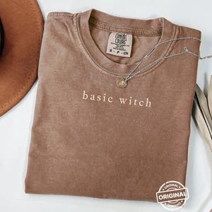 May include: A brown Comfort Colors t-shirt with the words "basic witch" printed in white. A silver necklace with a small pendant rests on the shirt. The shirt is folded and has a vintage look.