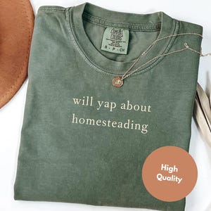 K&ouml;nnte beinhalten: Olivgr&uuml;nes T-Shirt mit dem wei&szlig;en Schriftzug &bdquo;will yap about homesteading&ldquo;. Eine goldfarbene Halskette mit Anh&auml;nger liegt auf dem Shirt. Ein brauner Hut ist im Hintergrund zu sehen. Ein runder Aufkleber mit &bdquo;High Quality&ldquo; befindet sich auf dem Shirt.