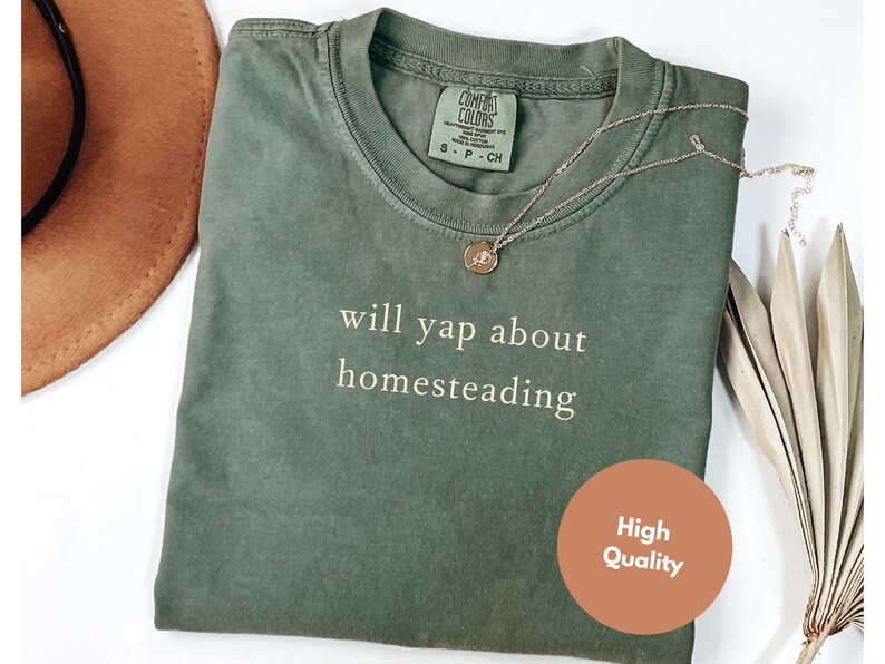 K&ouml;nnte beinhalten: Olivgr&uuml;nes T-Shirt mit dem wei&szlig;en Schriftzug &bdquo;will yap about homesteading&ldquo;. Eine goldfarbene Halskette mit Anh&auml;nger liegt auf dem Shirt. Ein brauner Hut ist im Hintergrund zu sehen. Ein runder Aufkleber mit &bdquo;High Quality&ldquo; befindet sich auf dem Shirt.