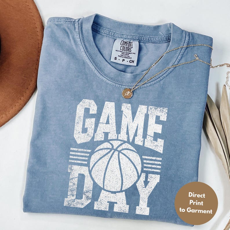 Puede incluir: Una camiseta azul Comfort Colors con un gr&aacute;fico de baloncesto "GAME DAY" en blanco. La camiseta tiene cuello redondo y mangas cortas. Un collar dorado con un colgante est&aacute; drapeado sobre la camiseta. La camiseta est&aacute; doblada sobre una superficie blanca.