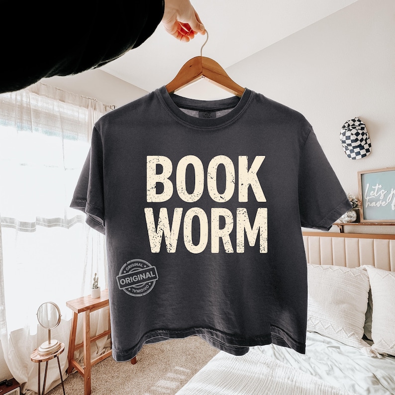 May include: A dark gray t-shirt with the words "BOOK WORM" printed in a distressed, off-white font. The shirt is hanging on a wooden hanger. A small, circular "ORIGINAL" stamp is visible near the bottom of the shirt.