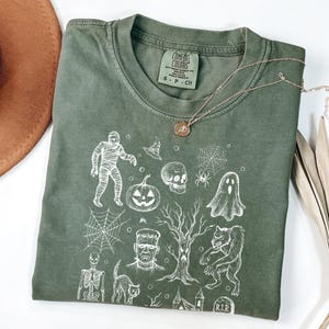 May include: An olive green Comfort Colors t-shirt with a white Halloween graphic. The design includes a mummy, pumpkin, skull, spiderwebs, and a werewolf. A silver necklace with a pendant is also visible.
