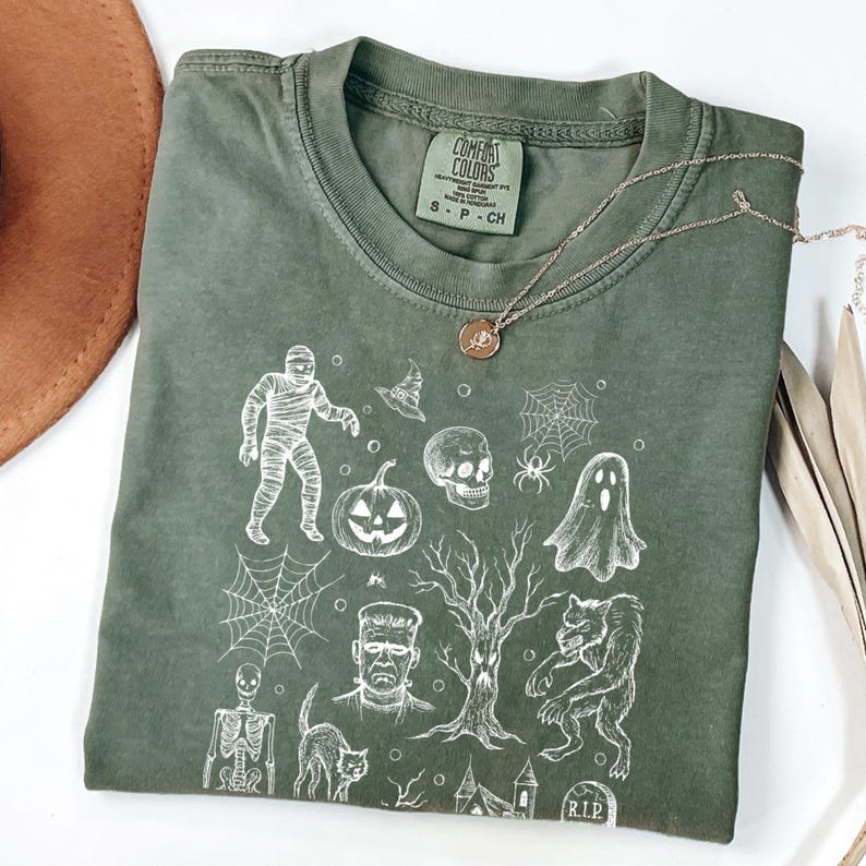 May include: An olive green Comfort Colors t-shirt with a white Halloween graphic. The design includes a mummy, pumpkin, skull, spiderwebs, and a werewolf. A silver necklace with a pendant is also visible.