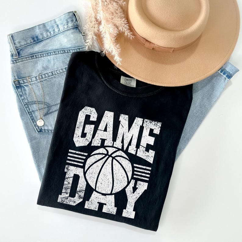 Puede incluir: Una camiseta negra con las palabras "GAME DAY" y un gr&aacute;fico de baloncesto en blanco. La camiseta est&aacute; colocada sobre unos vaqueros azul claro, con un sombrero color canela y un collar visible. Tambi&eacute;n hay una ramita de flores secas.
